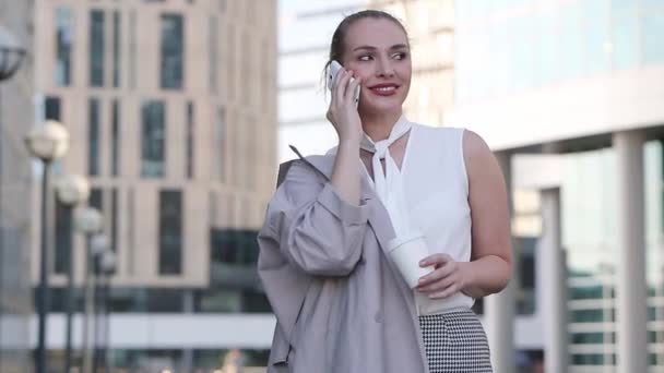 Jovem mulher de negócios com xícara de café falando no smartphone em pé entre o centro da cidade — Vídeo de Stock