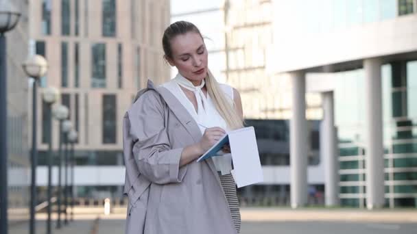 Joven mujer de negocios escribiendo en su cuaderno de pie entre el centro de la ciudad — Vídeo de stock