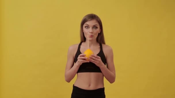 Sportliches Mädchen trinkt Orangensaft mit einem Cocktail-Strohhalm. Studio, gelber Hintergrund — Stockvideo