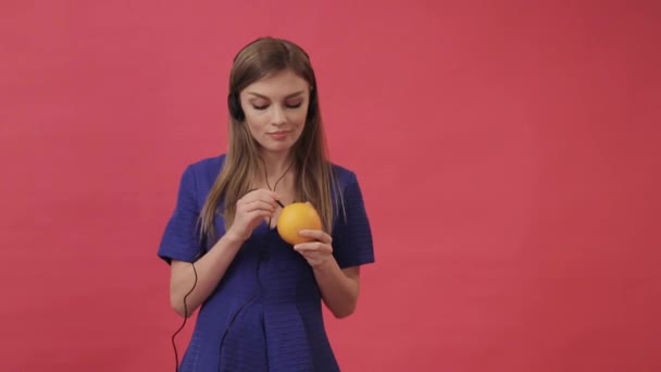 Красива дівчина в сукні з'єднує навушники з апельсином, а потім танцює і слухає музику. Студія, фіолетовий фон — стокове відео