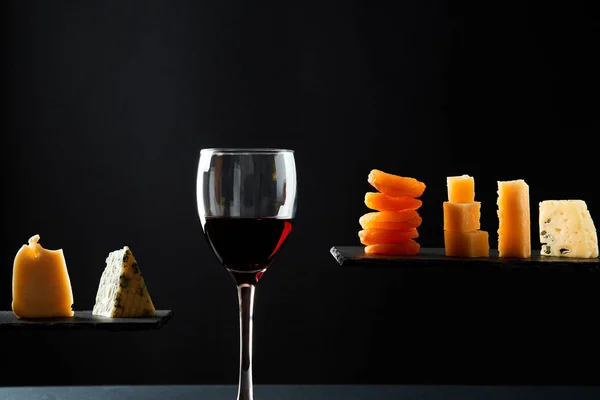 Trozos de queso y fruta seca al lado de un vaso de vino tinto dulce sobre fondo negro — Foto de Stock