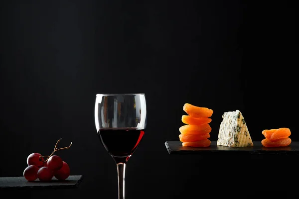 Stukjes blauwe kaas en gedroogd fruit naast glas rode zoete wijn op zwarte achtergrond — Stockfoto