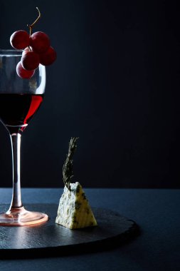 Siyah arka planda üzüm demeti ve mavi peynir parçası ile süslenmiş lezzetli kırmızı şarap kadehi.