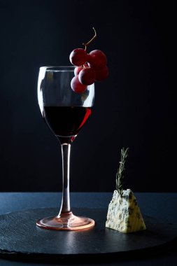 Siyah arka planda üzüm demeti ve mavi peynir parçası ile süslenmiş lezzetli kırmızı şarap kadehi.