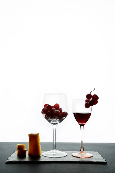 Een paar stukjes kaas en cherry tomaten naast een glas rode wijn gevuld met druiven op een witte achtergrond — Stockfoto