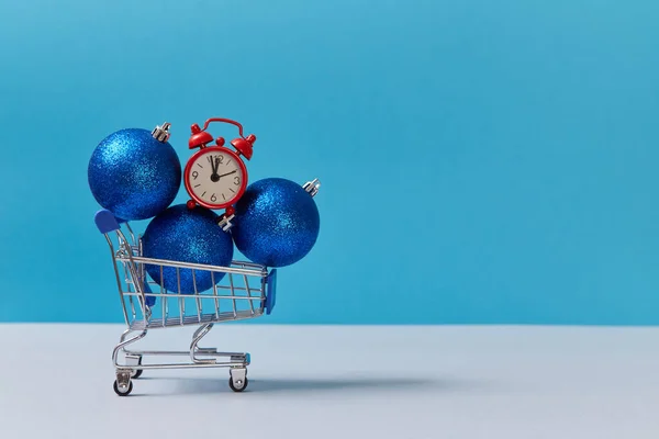 Carro de compras con bolas de purpurina y reloj rojo. Doce en punto. Año nuevo y Navidad. Copiar espacio. Aislado sobre fondo azul —  Fotos de Stock