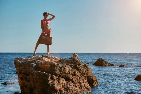 Adventure trip idea. Lady in pink bikini with suitcase standing on rock. Summer holiday. Time for travel. Beach vacation — Stock Photo, Image