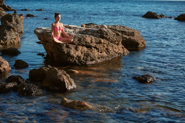 Nyugodt hölgy rózsaszín bikiniben meditál a sziklán. Nyugodt tenger. Egyedül voltam. Meditáció és önimádat. Szabadidő a szabadban — Stock Fotó