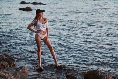 Plaj tatili Dinlenme ve akşam yürüyüşü zamanı. Deniz dalgalarında küçük kayalarda poz veren zinde bir kadın. Güzel doğa manzarası