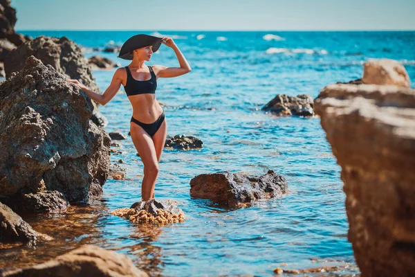 Γυναίκα με μαύρο μαγιό και καπέλο που ποζάρει στη θάλασσα. Παραλία διακοπές και το καλοκαίρι έννοια ώρα, αντιγραφή χώρου. Καταπληκτική θέα στη θάλασσα — Φωτογραφία Αρχείου