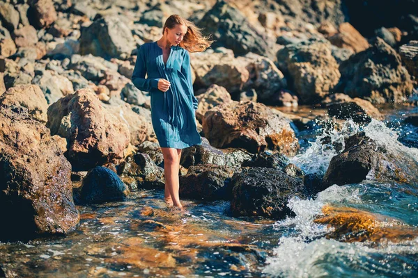 Een vrouw in een blauwe jurk die over de kustlijn loopt. Scherp rotsachtig terrein. Meditatie en gezondheid. Zeegezicht en ontspanning concept — Stockfoto