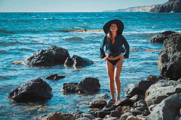 Glimlachende en gelukkige vrouw met hoed. Rotsachtige kustlijn en blauwe zee. Meditatie en moment van ontspanning. Zomervakantie — Stockfoto