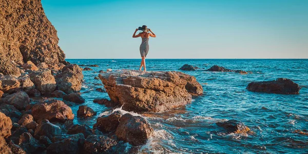 Γυναίκα ποζάρει με μοντέρνο καπέλο. Θέα στο βουνό και στη θάλασσα. Βραχώδης ακτογραμμή και παραλία έννοια διακοπών. Φύση και χρόνος για χαλάρωση — Φωτογραφία Αρχείου