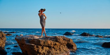 Keskin kayalıklarda poz veren tatlı bir kadın. Moda şapkalı bir bayan. Nefes kesici deniz manzarası. Plaj ve rahatlama konsepti, kopyalama alanı
