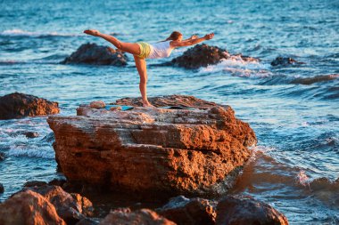 Sportif bir kadın tek ayak üzerinde dengede duruyor. Okyanus manzarası ve temiz hava. Formda vücut için pilates. Esneme süresi ve yoga treni konsepti