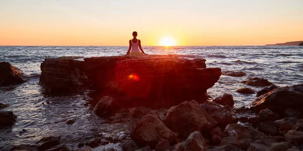 Όμορφο ηλιοβασίλεμα και θέα στον ωκεανό. Η γυναίκα διαλογίζεται στην πέτρα. Ώρα για σκέψη και γιόγκα. Πιλάτες και τέντωμα — Φωτογραφία Αρχείου