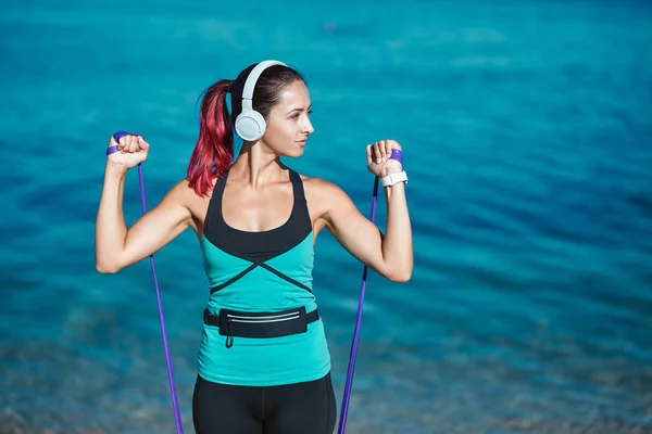 Brazos de entrenamiento de señora con bandas elásticas y escuchar música en auriculares. Océano azul sobre fondo. Deporte y fitness idea — Foto de Stock