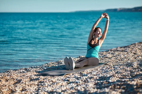 Mosolygó nő nyújtózkodik a jóga szőnyegen ülve. Gyönyörű kilátás a tengerre és a friss levegőre. Egészséges életmód és sport koncepció — Stock Fotó