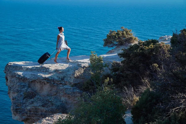 Concepto de viaje y trabajo remoto. Vida trabajo equilibrio idea. Mujer joven con maleta y portátil que va en roca de mar. —  Fotos de Stock