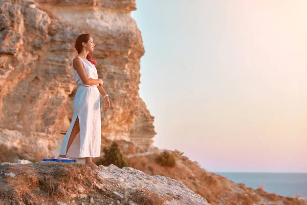 Χαριτωμένη κυρία με λευκό φόρεμα που ποζάρει στο βράχο. Ηλιοβασίλεμα στη θάλασσα και τη φύση. Διαμόρφωση και αυτο-απορροφούμενη ιδέα, αντιγραφή χώρου — Φωτογραφία Αρχείου