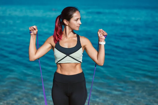 Brunette lady posing with rubber bands. Training on fresh air near ocean view. Fitness and healthy lifestyle concept