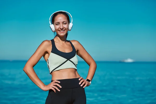 Hermosa chica posando en ropa deportiva cómoda. La señora escucha música en los auriculares. Vista al mar sobre telón de fondo. Deporte y naturaleza idea — Foto de Stock