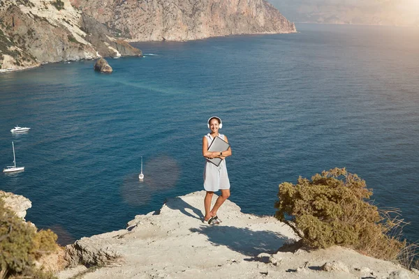 Femme en vacances d'été. Dame avec ordinateur portable et casque. Vue océan et nature vierge. Bureau à distance et idée de temps de voyage — Photo