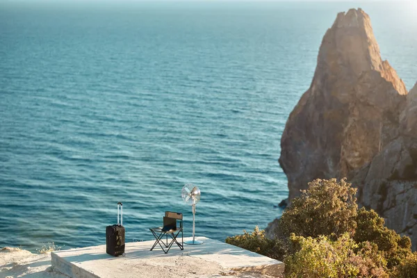 Chaise touristique et ordinateur portable, valise et ventilateur sur rocher de mer. Paysage marin sur fond. Travail à distance et idée de changement de style de vie — Photo