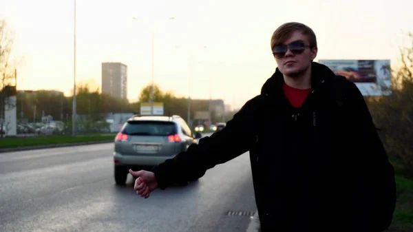 El hombre atractivo está tomando un taxi cerca de la carretera. Un joven feliz coge un taxi en las calles europeas. Retrato de un turista caucásico con mochila sonriendo y cogiendo un taxi — Foto de Stock