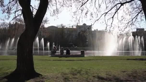 Городской фонтан Запись. Люди гуляют по парку. Фонтан в городском парке в жаркий летний день. Поток воды, капли и яркие брызги воды в красивом городском фонтане — стоковое видео