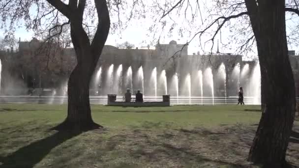 Fonte da cidade. Filmagem. Pessoas a andar no parque. Fonte no parque da cidade no dia quente do verão. Fluxo de água, gotas e salpicos brilhantes de água na bela fonte da cidade — Vídeo de Stock