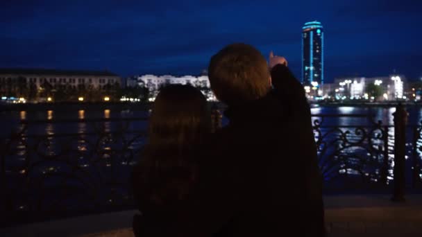 Pareja joven observando un paisaje urbano de pie en un muelle de un río. Pareja joven cerca del río en la ciudad por la noche — Vídeos de Stock