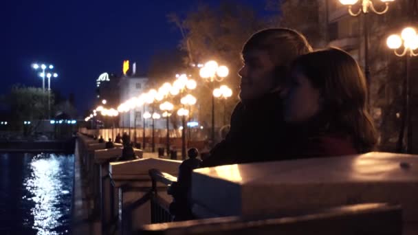 Jong koppel kijken een stadsgezicht staande op een kade van een rivier. Jong koppel in de buurt van de rivier in de stad bij nacht — Stockvideo
