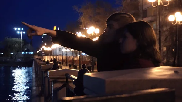 Pareja joven observando un paisaje urbano de pie en un muelle de un río. Pareja joven cerca del río en la ciudad por la noche — Foto de Stock