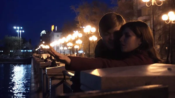 Pareja joven observando un paisaje urbano de pie en un muelle de un río. Pareja joven cerca del río en la ciudad por la noche — Foto de Stock