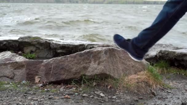 Primo piano della gamba di un uomo che indossa jeans in piedi su una roccia nel fiume. Le scarpe da ginnastica calpestano una pietra. Chiudere il piede vicino all'acqua in una giornata tempestosa — Video Stock