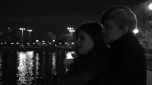 A couple in love stands against the lights of a night city at waterfront. Amazing wedding couple near the river at night, monochrome — Stock Photo, Image