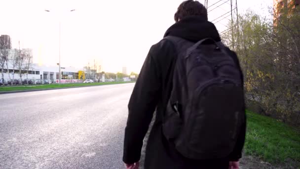 Man met een rugzak wandelen langs de weg. Bebaarde hipster met rugzak en jeans jas en jeans wandelen gaat met zijn rug langs een asfaltweg platteland — Stockvideo