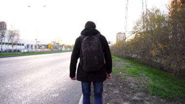 Adam yolda yürürken bir sırt çantası ile. Sırt çantası ve kot ceket ve kot pantolon yürüyüş ile sakallı hippi sırtını bir kırsal asfalt yol boyunca gider