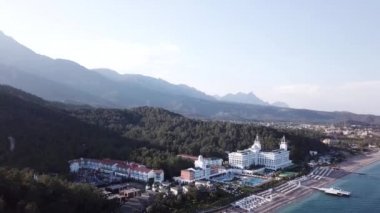 Havadan görünümü ile orman ve dağlar arka plan ve mavi gökyüzü ile lüks otel sahil üzerinde. Video. Beyaz lüks otelin dağlar, orman ve gökyüzü ile kıyı şeridi denizde güzel yaz üstten görünüm
