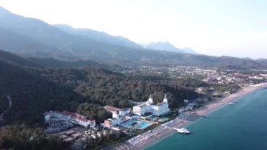 Havadan görünümü ile orman ve dağlar arka plan ve mavi gökyüzü ile lüks otel sahil üzerinde. Video. Beyaz lüks otelin dağlar, orman ve gökyüzü ile kıyı şeridi denizde güzel yaz üstten görünüm