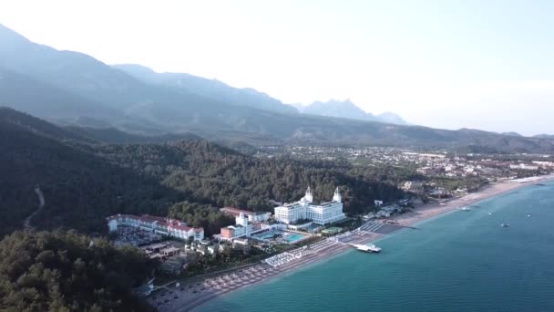 Vídeo. Vista aérea del hotel en la orilla del mar y las montañas cubiertas de bosque verde, árboles con cielo azul al amanecer. Vídeo. Vista aérea en la orilla del mar con hotel de lujo con fondo de bosque y montañas — Vídeos de Stock