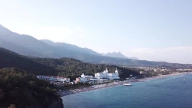 Havadan görünümü ile orman ve dağlar arka plan ve mavi gökyüzü ile lüks otel sahil üzerinde. Video. Beyaz lüks otelin dağlar, orman ve gökyüzü ile kıyı şeridi denizde güzel yaz üstten görünüm