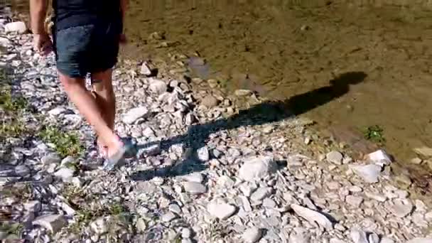 Boy walks the rocks in the water to the shore. Feet man walking outdoor Travel Lifestyle vacations concept with lake and sun on background. Boy tries to keep his balance when walking on the slippery — Stock Video