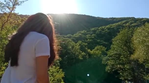 若いブルネットの少女は、立ちバック。山の背景。夕日を見て素晴らしい若い女性で背中側面図です。女の子の髪を開発風、太陽と山背景の背面図 — ストック動画