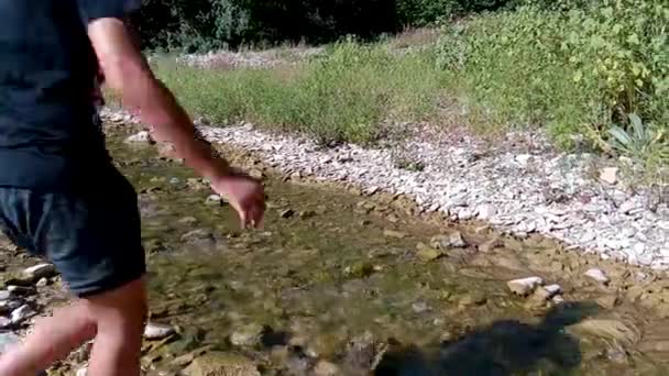 Fiú keresztezi a patak. láb fiú Vértes a háttérben a zubogó erdei forrás és sziklák. A koncepció a túrázás. Egy fiatal fiú sétált a folyó partján a kövek, egy napsütéses nyári napon — Stock videók
