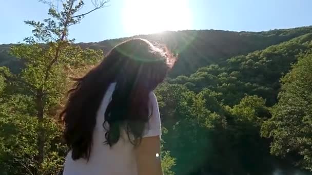 Νεαρή μελαχρινή κοπέλα στέκεται πίσω. Με φόντο βουνά. Πίσω πλευρά θέα σε μια υπέροχη νεαρή γυναίκα βλέποντας το ηλιοβασίλεμα. Πίσω όψη του κορίτσι μαλλιά αναπτύσσει ανέμου, φόντο βουνά με ήλιο — Αρχείο Βίντεο