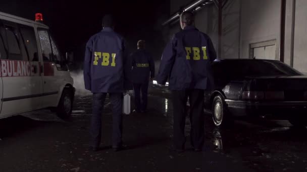 Gli agenti dell'FBI lavorano sulla scena di notte, auto della polizia con luci e sfondo ambulanza. Vista posteriore su tre agenti dell'FBI andare verso la scena del crimine — Video Stock