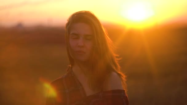 Primer plano retrato de una joven atractiva sonriendo al aire libre. Mujer en el fondo del atardecer del campo de la estepa en tiempo ventoso. Retrato de una mujer atractiva en la naturaleza al atardecer — Vídeo de stock