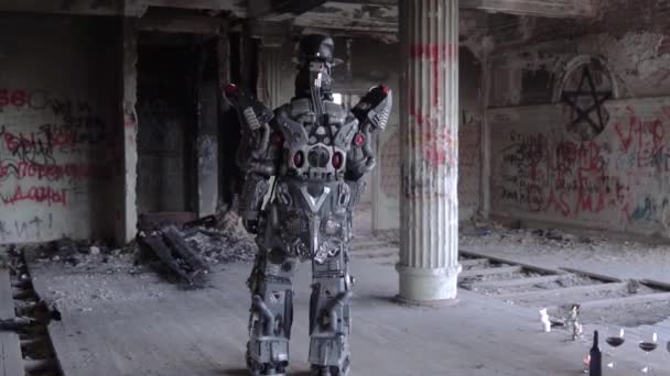 Humanoïde robot staat met zijn rug in hoed in verlaten gebouw. Beeldmateriaal. Android op date met glazen en een fles wijn staat terug tegen grijze muren met graffiti — Stockvideo